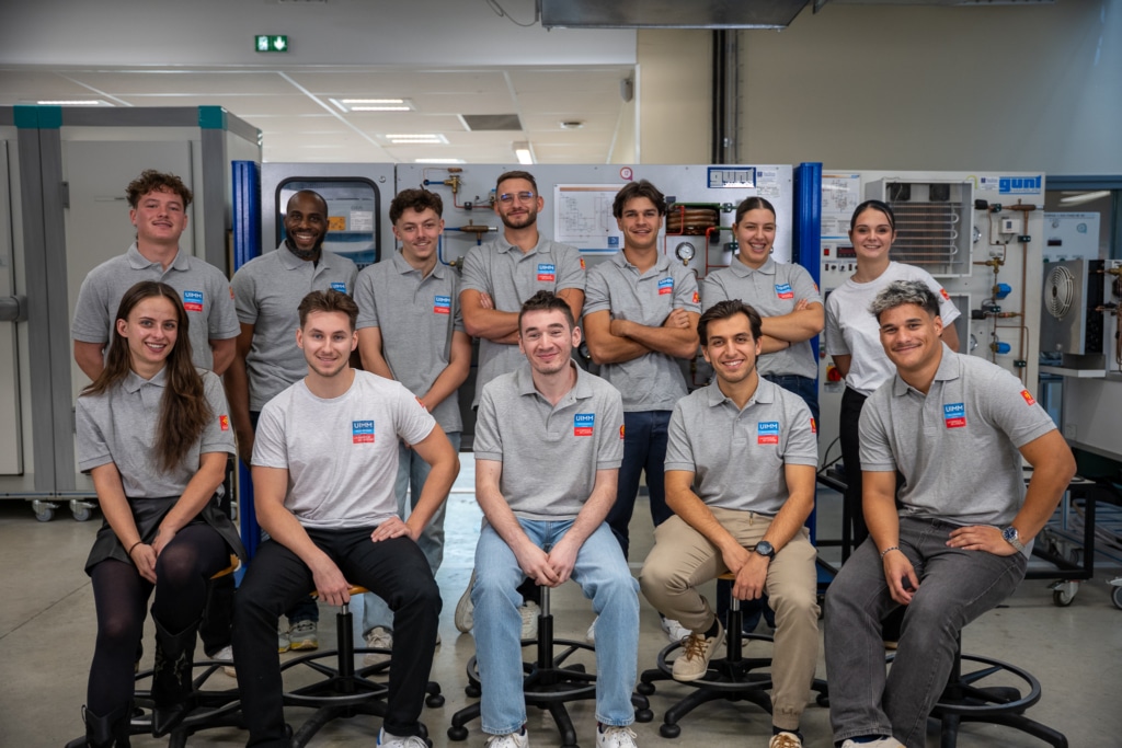 Group of eleven students in gray polo shirts posing in a technical workshop with machinery behind them, smiling at the camera beyond the stools.