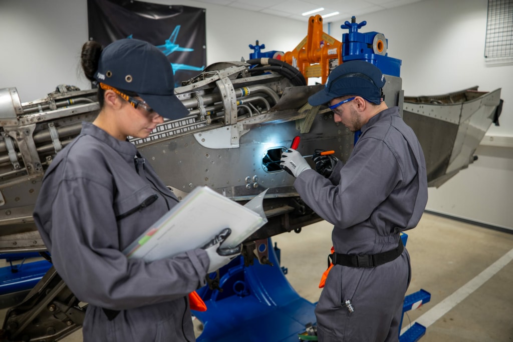 Two technicians in gray coveralls repair an aircraft fuselage in a maintenance hangar, using handheld tools and a light panel access.