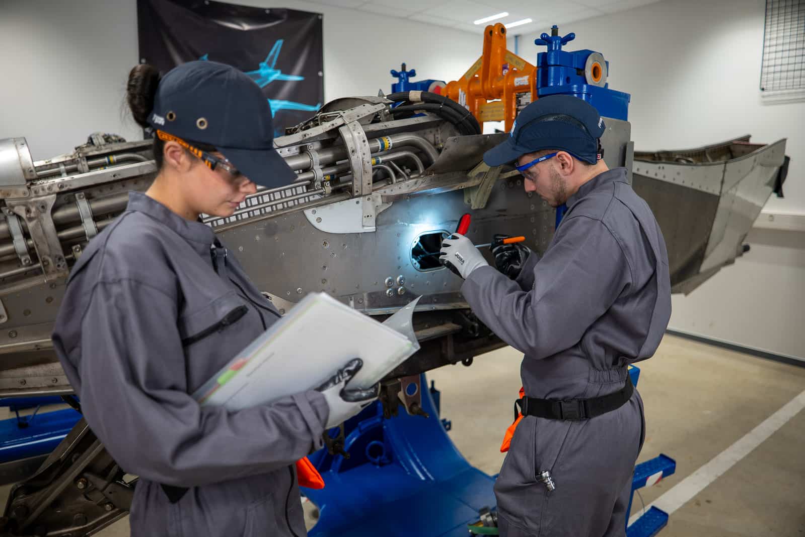Two technicians in gray coveralls repair an aircraft fuselage in a maintenance hangar, using handheld tools and a light panel access.