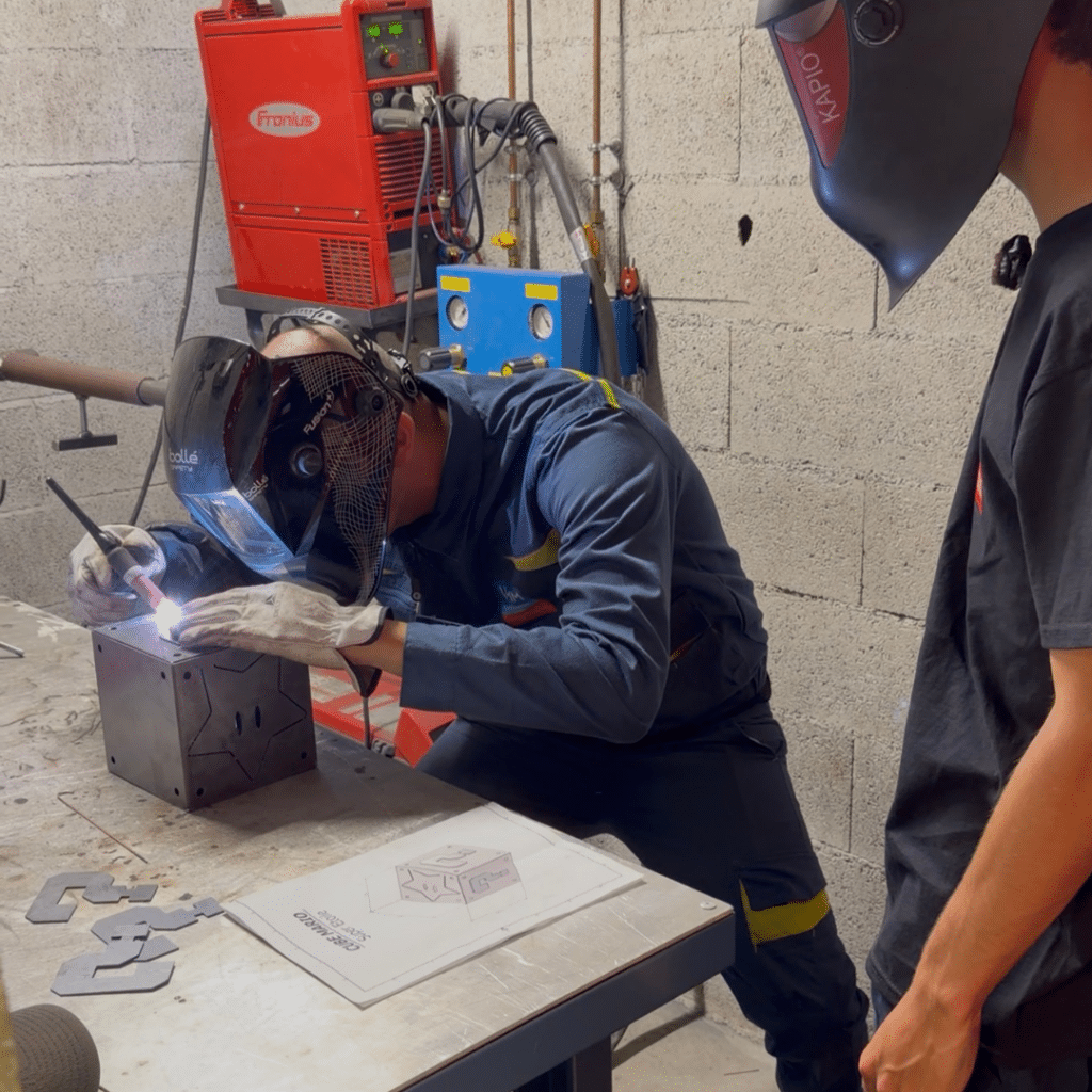 Welder in protective helmet and gloves welding a metal block on a workbench, sparks flying.