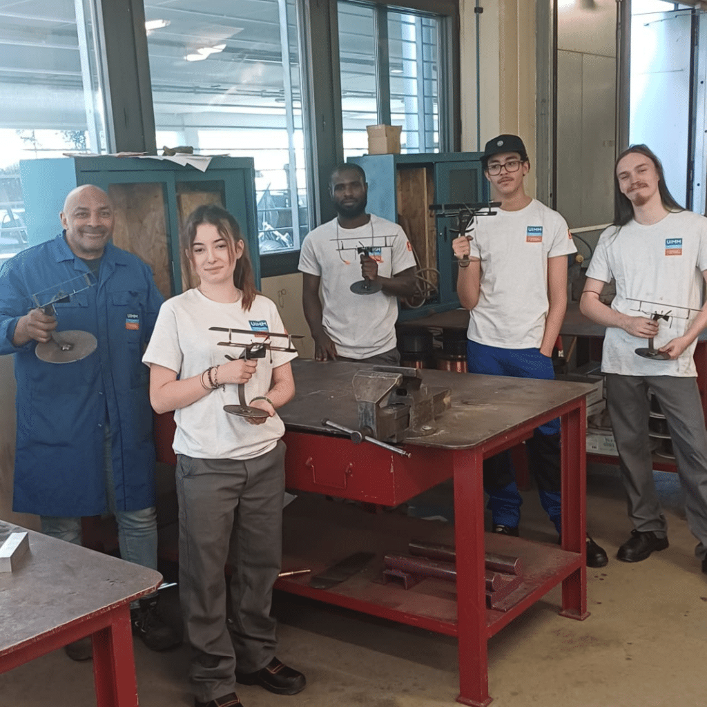 Five students and a man in a blue shop coat pose in a metalwork classroom, each holding a small metal stand or jig they built or are showcasing for a project.