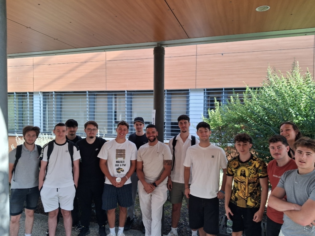 Group of young people posing for a photo outdoors in front of a wood-paneled building, some wearing backpacks and casual clothing, with greenery on the right.