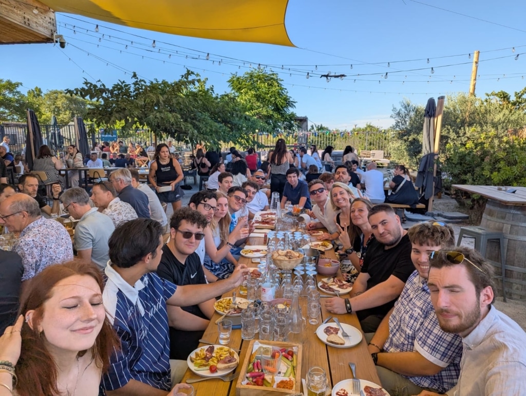 Large outdoor group picnic at a long wooden table, people eating and chatting under string lights.