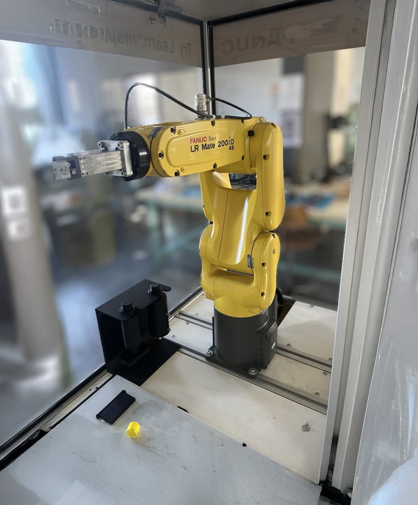 Yellow FANUC LR Mate robotic arm inside a glass enclosure, with a gripper at the end effector ready for manipulation in an industrial workstation.
