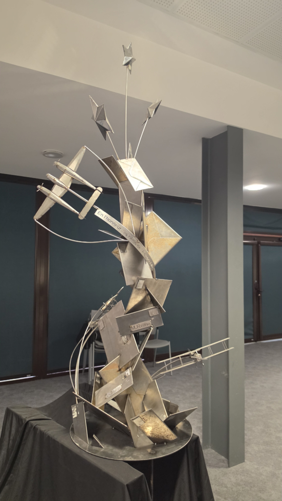 Abstract metal sculpture with geometric shapes on a pedestal in an indoor gallery.