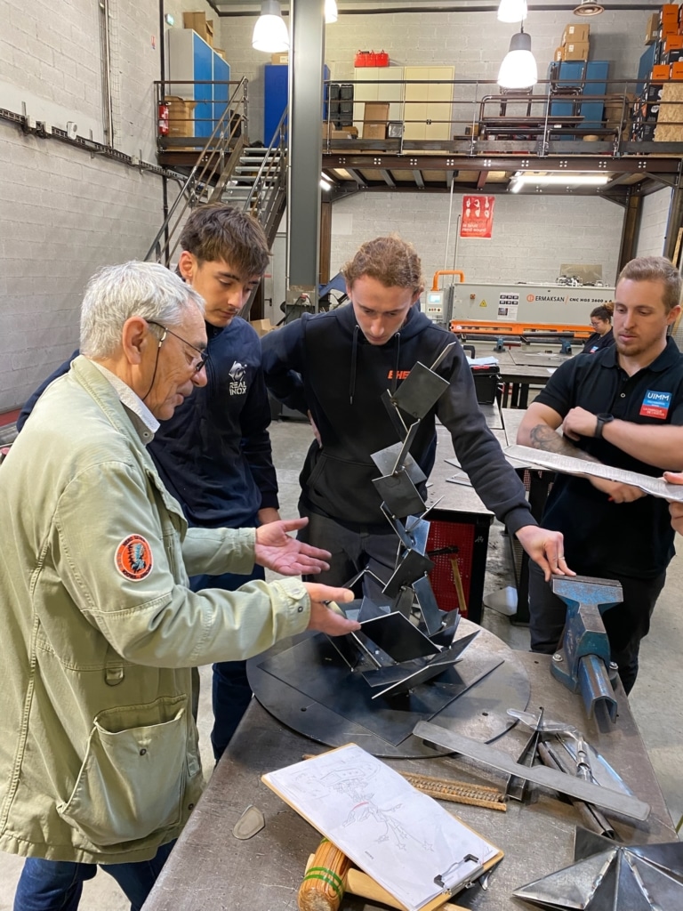 A group of four people studying metal pieces arranged on a workshop table, with blue cabinets and machinery in the background.