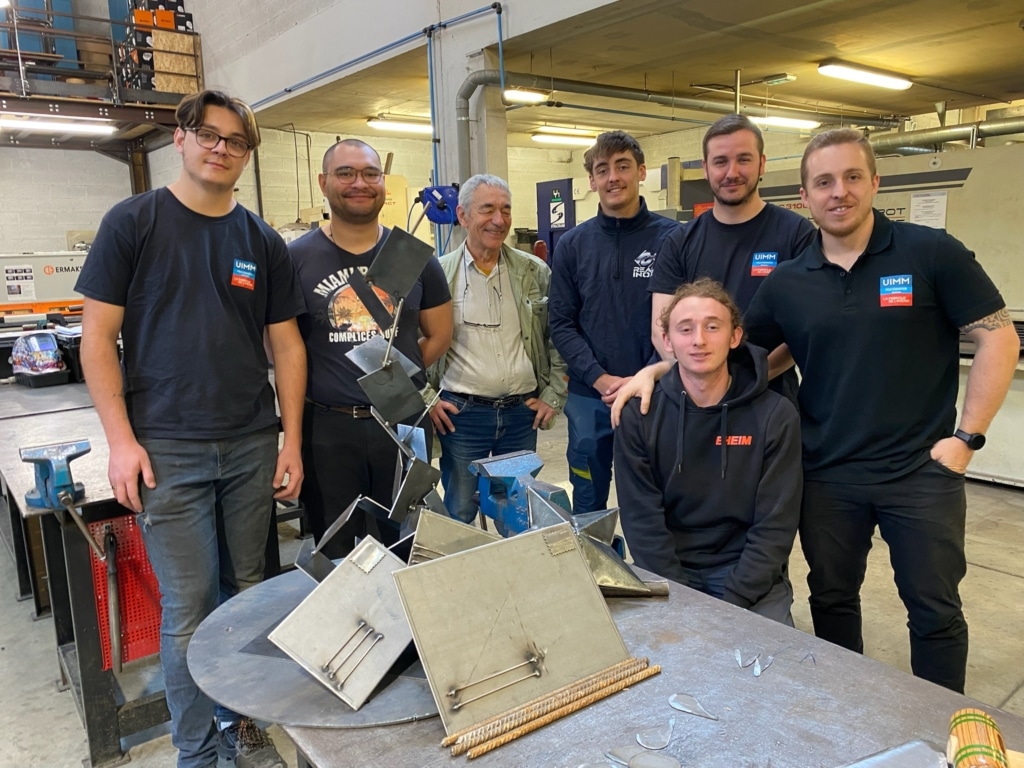 Group of six people in a metal workshop posing for the camera behind a table with metal fabrication pieces and tools visible in the foreground.