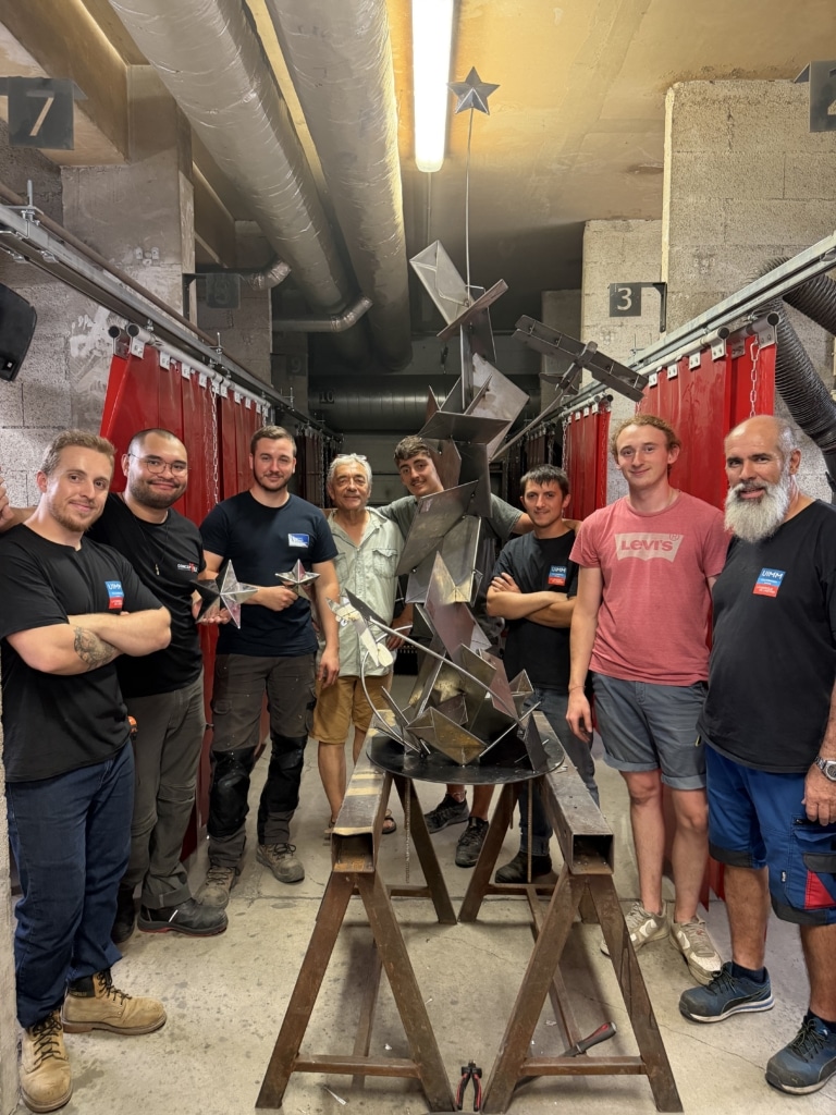 Group of men poses with a tall geometric metal sculpture in a workshop, with red curtains and industrial ducts above.