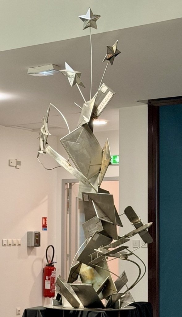 Abstract metal sculpture featuring angular, stacked squares and star-shaped elements rising from a pedestal in an indoor gallery setting.