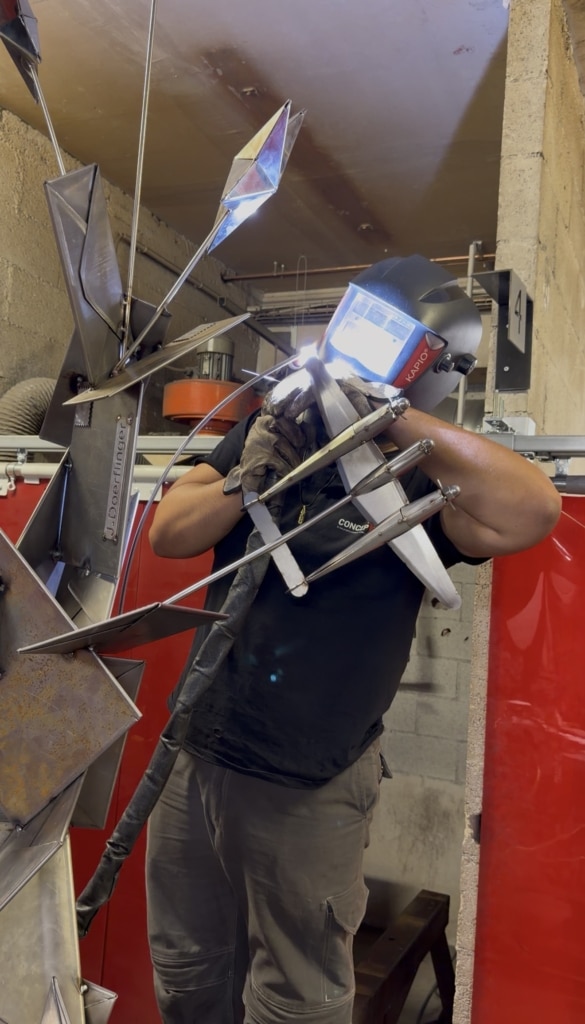 Welder in protective helmet and gloves assembling a sharp, angular metal sculpture in a workshop.