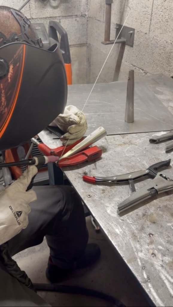 Welder in protective helmet and gloves at a metal workbench, feeding welding wire to a joint on metal tubing edges.