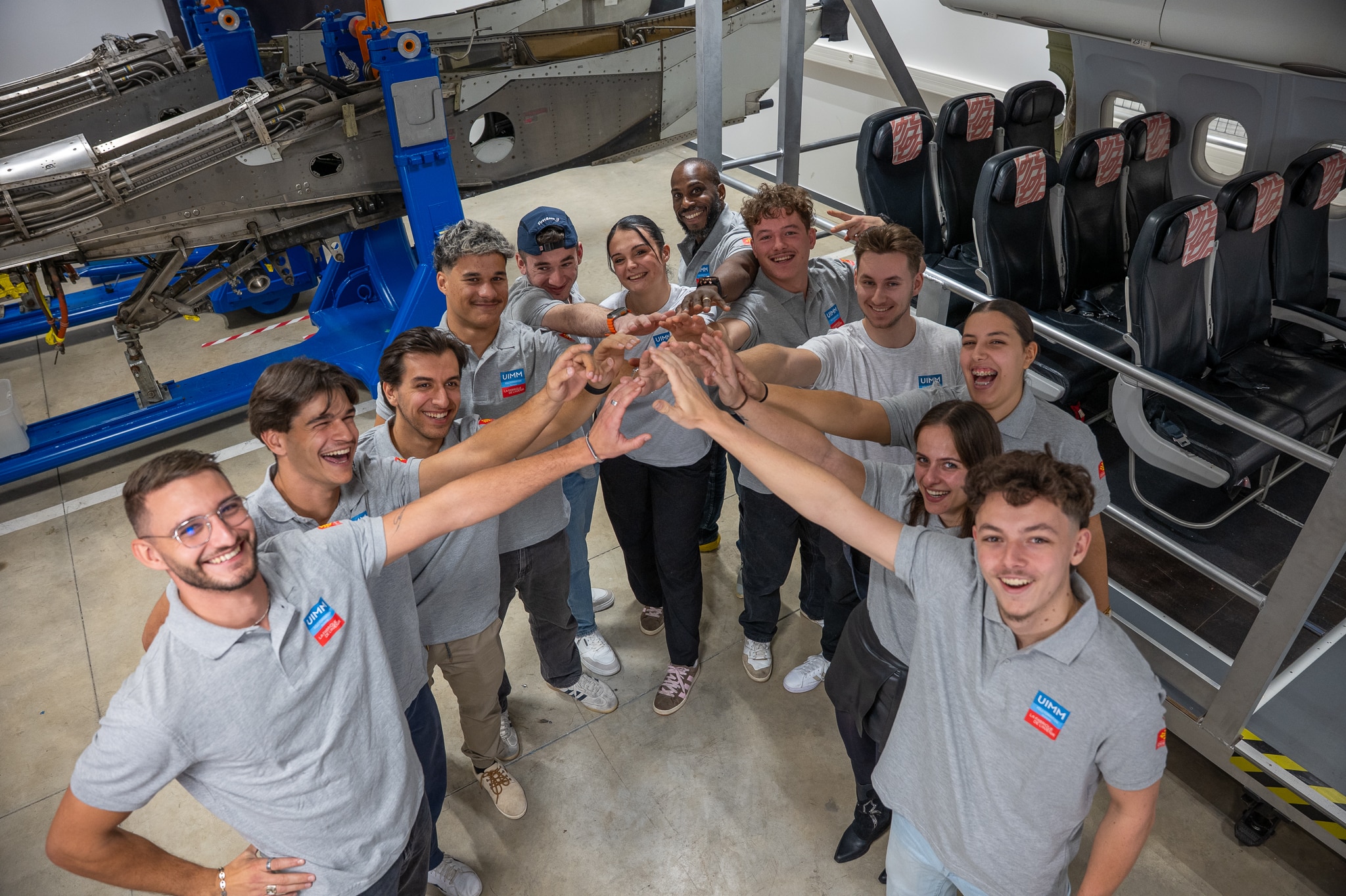A team of people in matching gray shirts forming a circle with hands joined in the center, smiling in a workshop with aircraft machinery and airplane seats in the background.