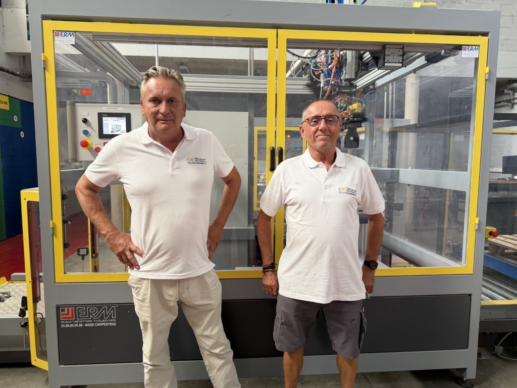 Two men in white polo shirts pose in front of a large yellow-framed industrial machine with glass panels in a factory setting.